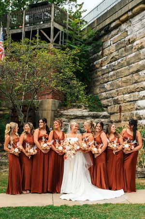 Bride with bridesmaids in rust orange gowns holding bouquets near stone wall outdoors.