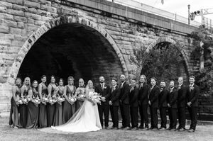 Large wedding party poses under stone arch bridge in black and white photo with bridesmaids and groomsmen.