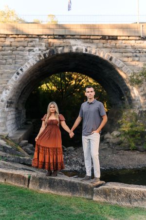 Couple holding hands in front of a stone arch bridge, woman in rust boho dress, man in gray shirt and khakis.
