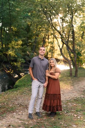 A couple poses hand-in-hand on a wooded path, woman in rust maxi dress, man in gray polo and khakis.