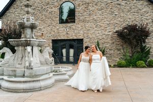Two brides in white strapless gowns standing together near stone fountain outside elegant wedding venue.