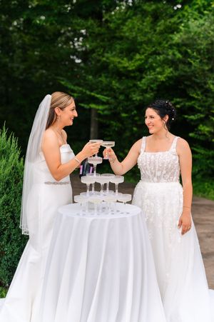 Two brides in white gowns toasting champagne glasses at an outdoor wedding celebration with a champagne tower.