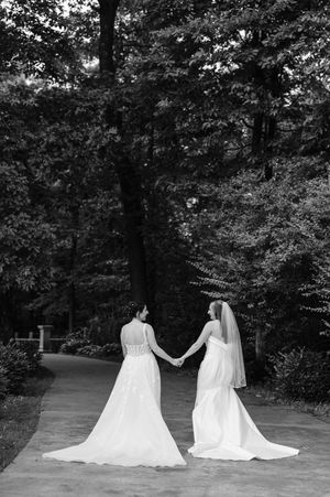 Two brides in white wedding gowns holding hands on a tree-lined path, black and white.