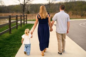 Family of three walking hand-in-hand on a park path, parents and young child viewed from behind.