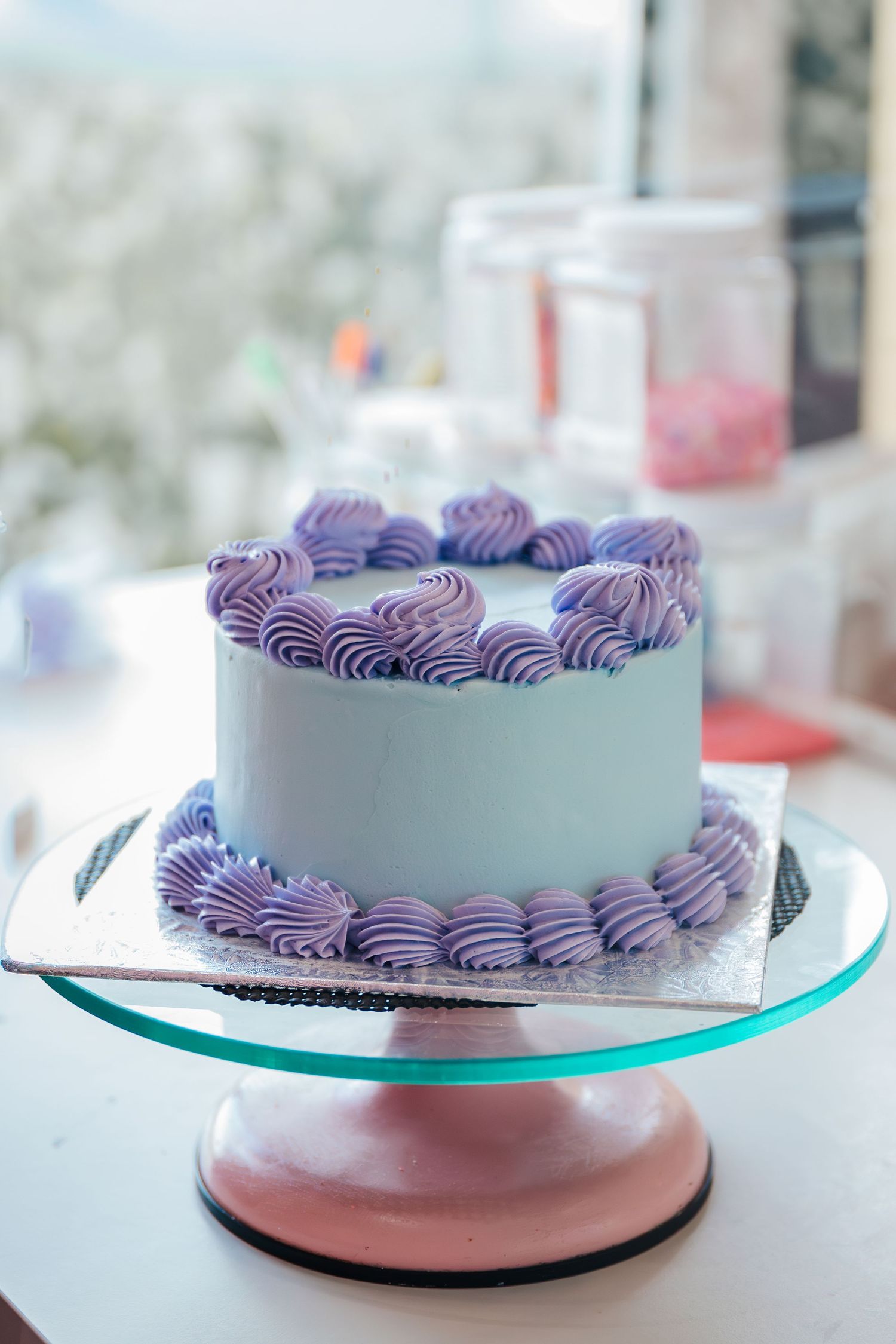 Blue cake with purple swirl rosette decorations on top and base, displayed on a pink glass cake stand. Bakery branding pictures.