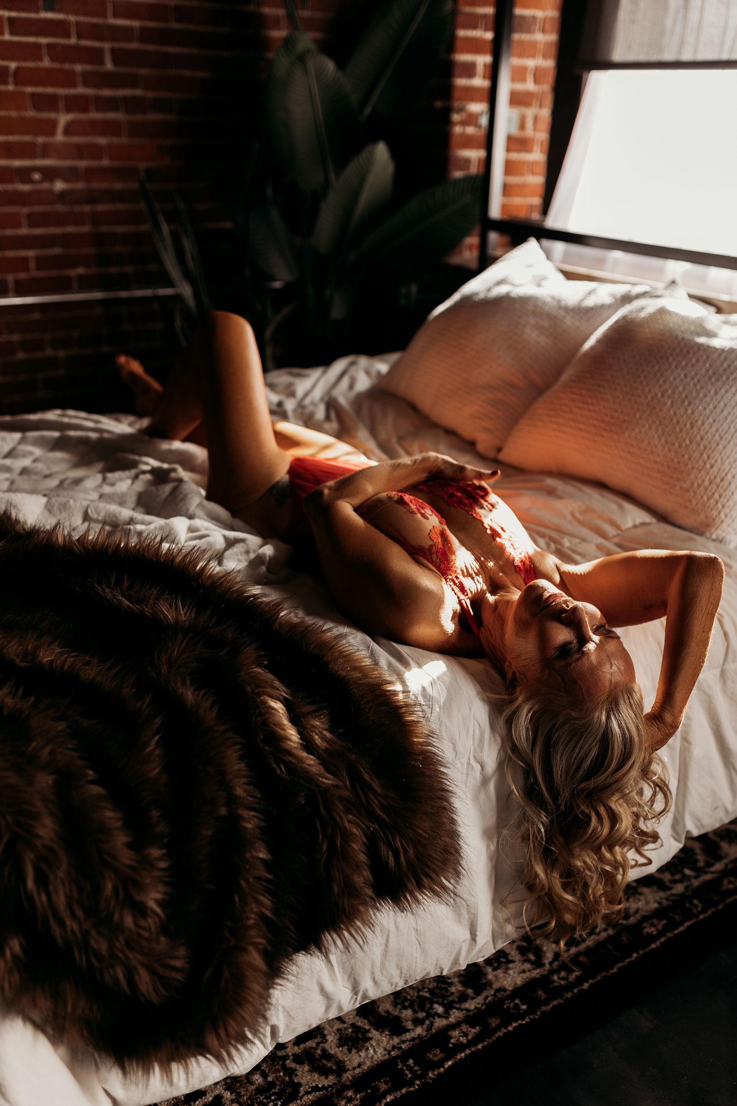 Woman in black lace lingerie lying on red satin sheets, back view showing corset detail and tattoos.
