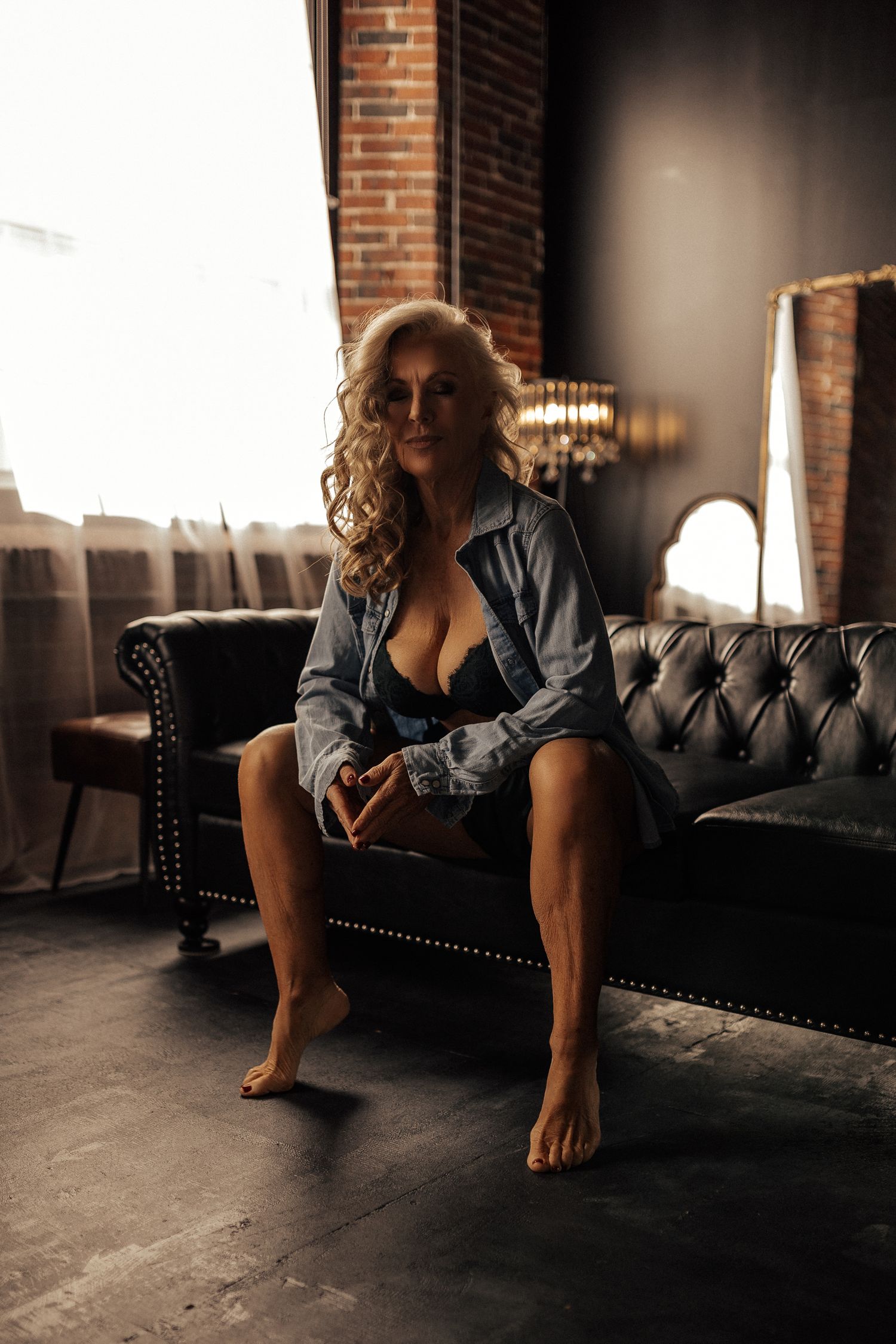 Woman in denim shirt and lingerie sitting on a tufted leather sofa in a moody loft-style interior.