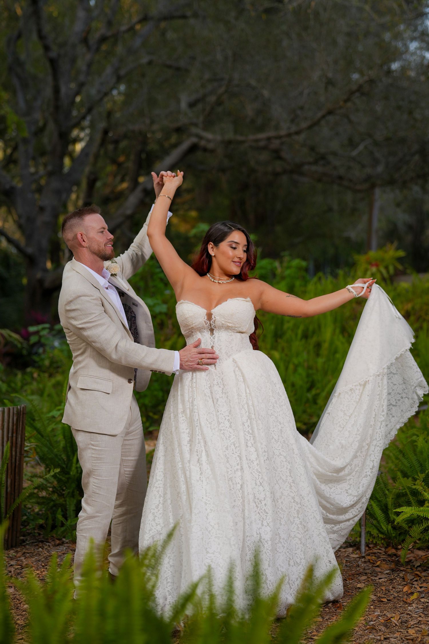 Bride in lace gown twirls with groom in tan suit during outdoor wedding portrait in lush garden setting.