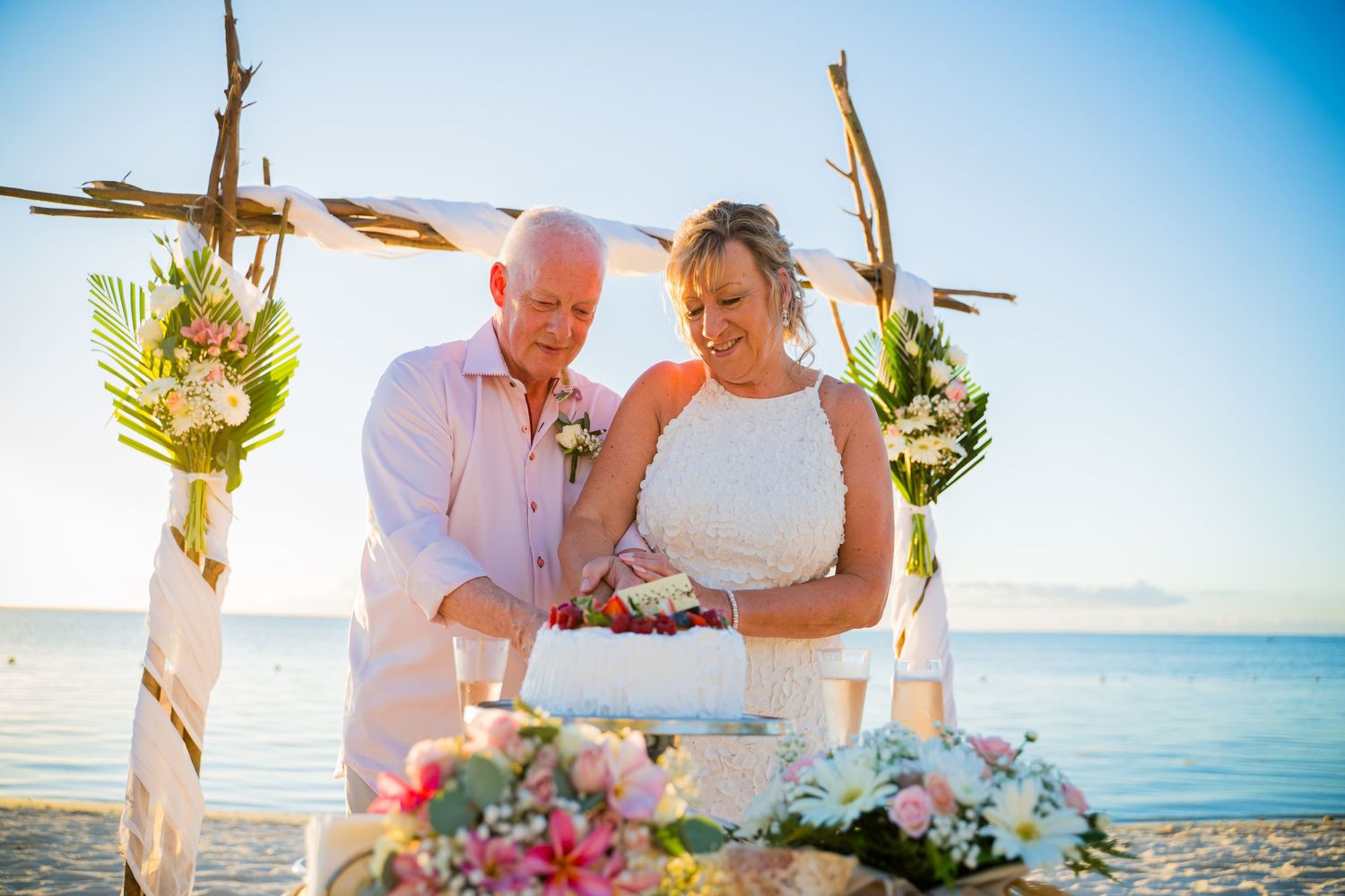 Julie and Stuart cutting their wedding cake under a driftwood arch on the beach in Mauritius