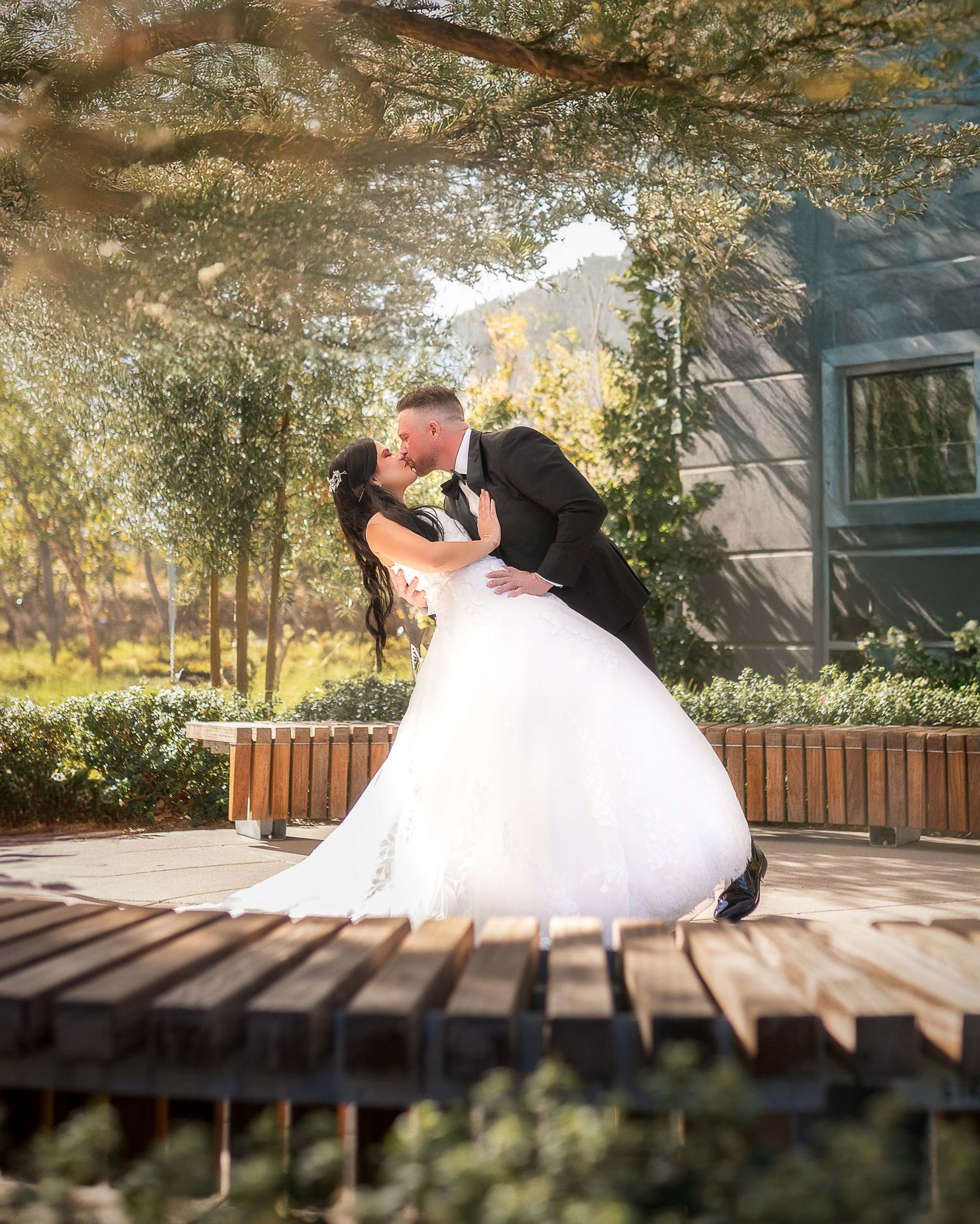 Bride in white ball gown and groom in black tux share a romantic dip kiss outdoors at a sunlit wedding venue.