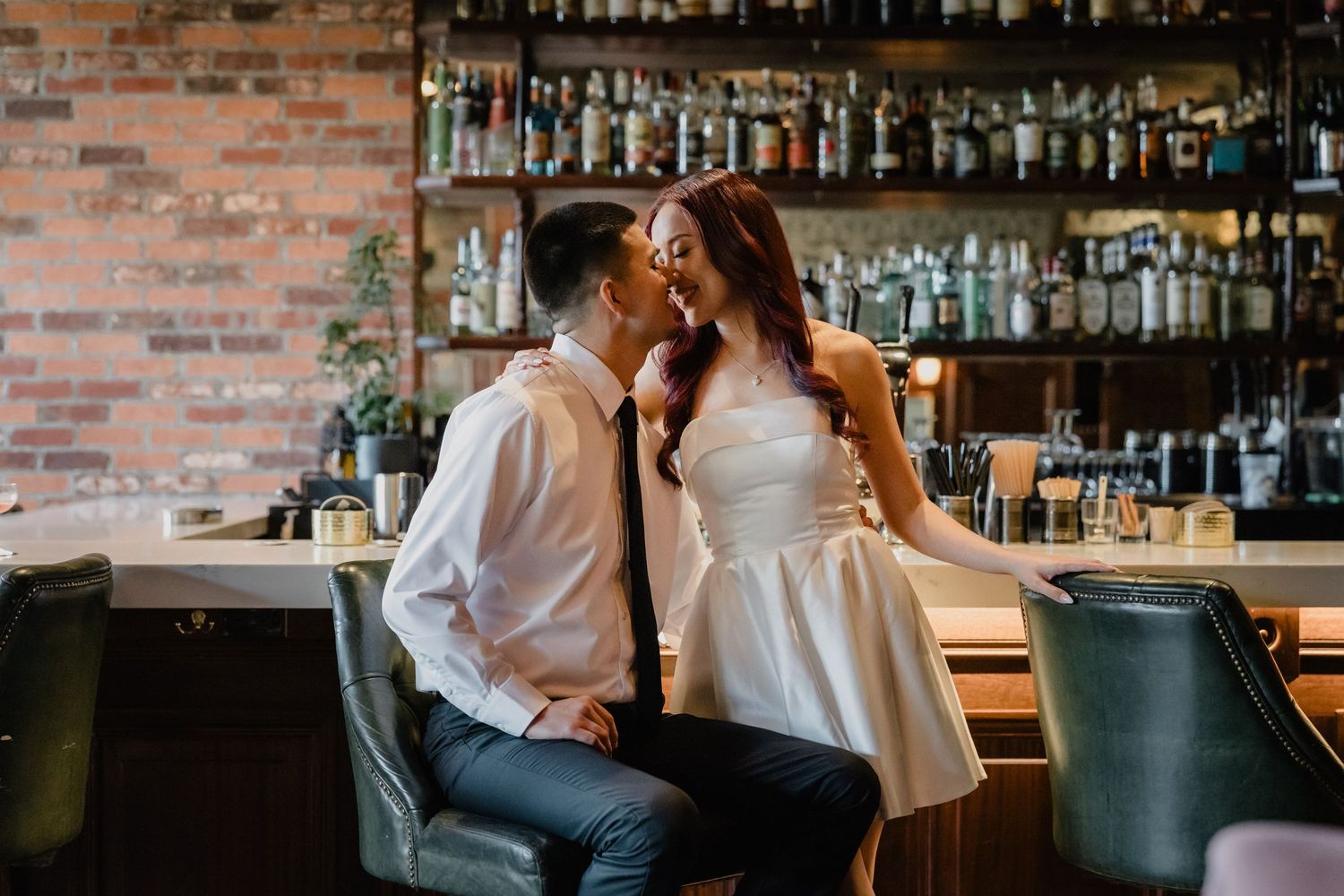 A wedding couple shares a romantic moment under a white umbrella in a rainy garden with flowers and a building in the background.