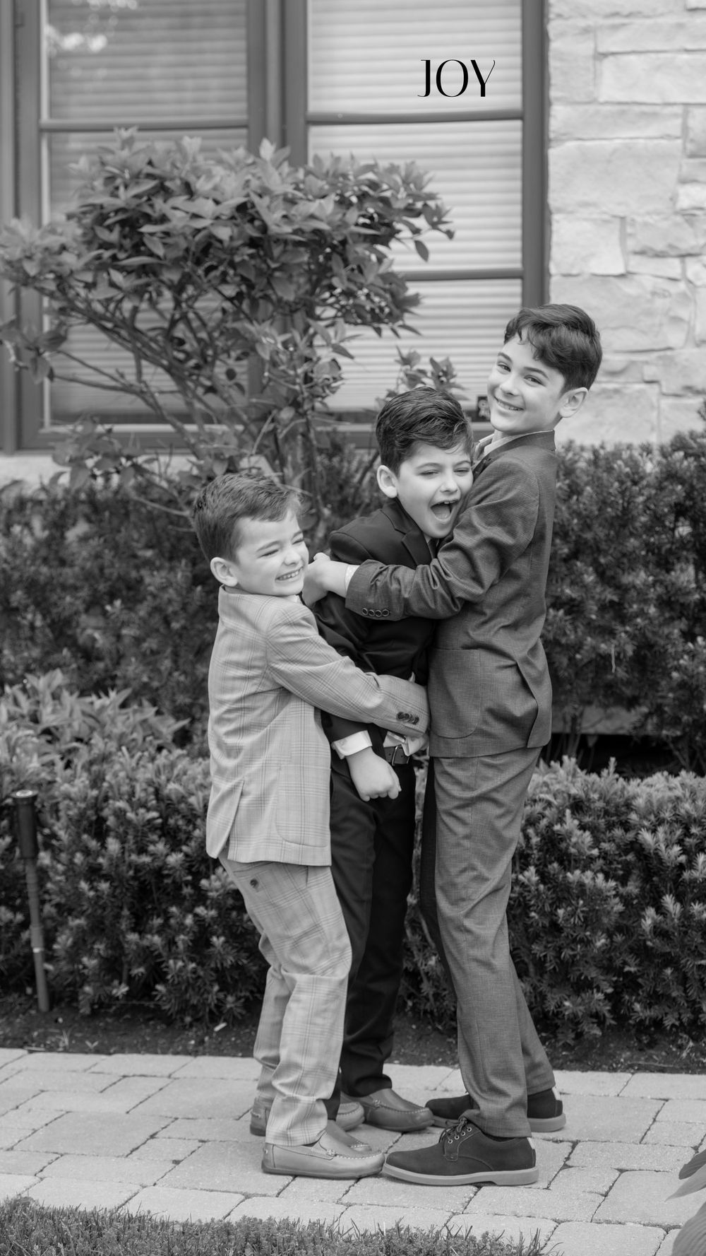 Three young boys in suits joyfully hugging outdoors in a black and white photo.