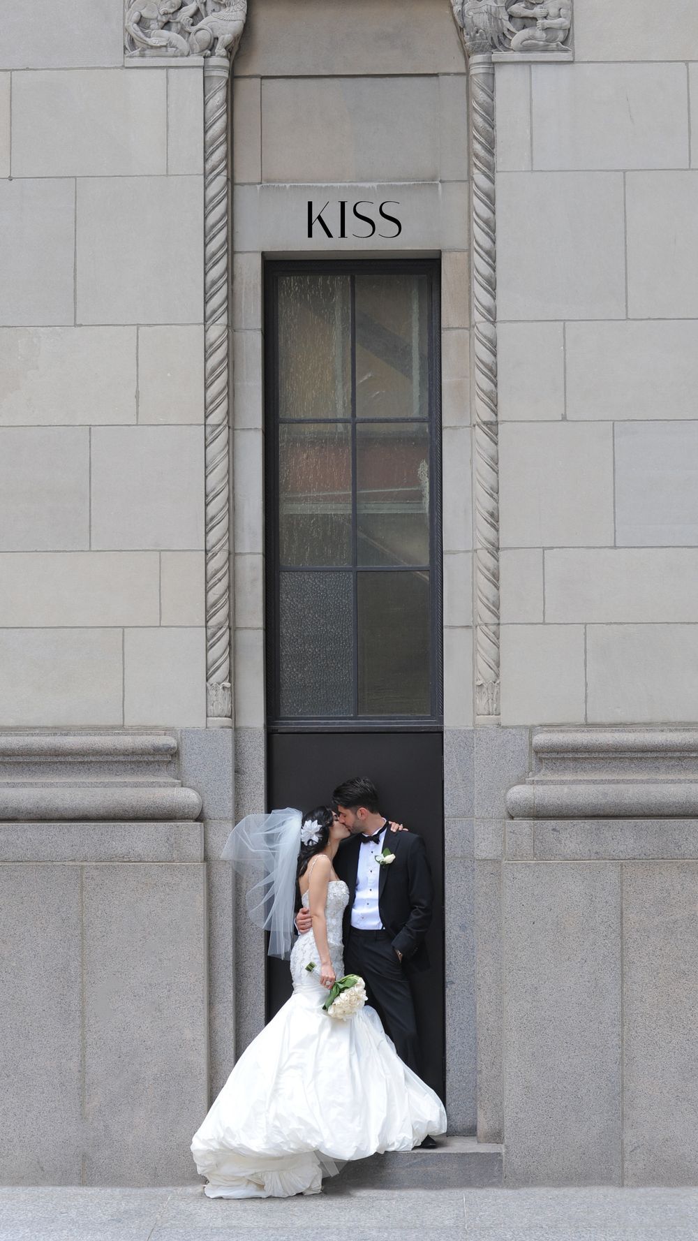 Bride and groom sharing a romantic kiss in a stone doorway labeled KISS during their wedding day.