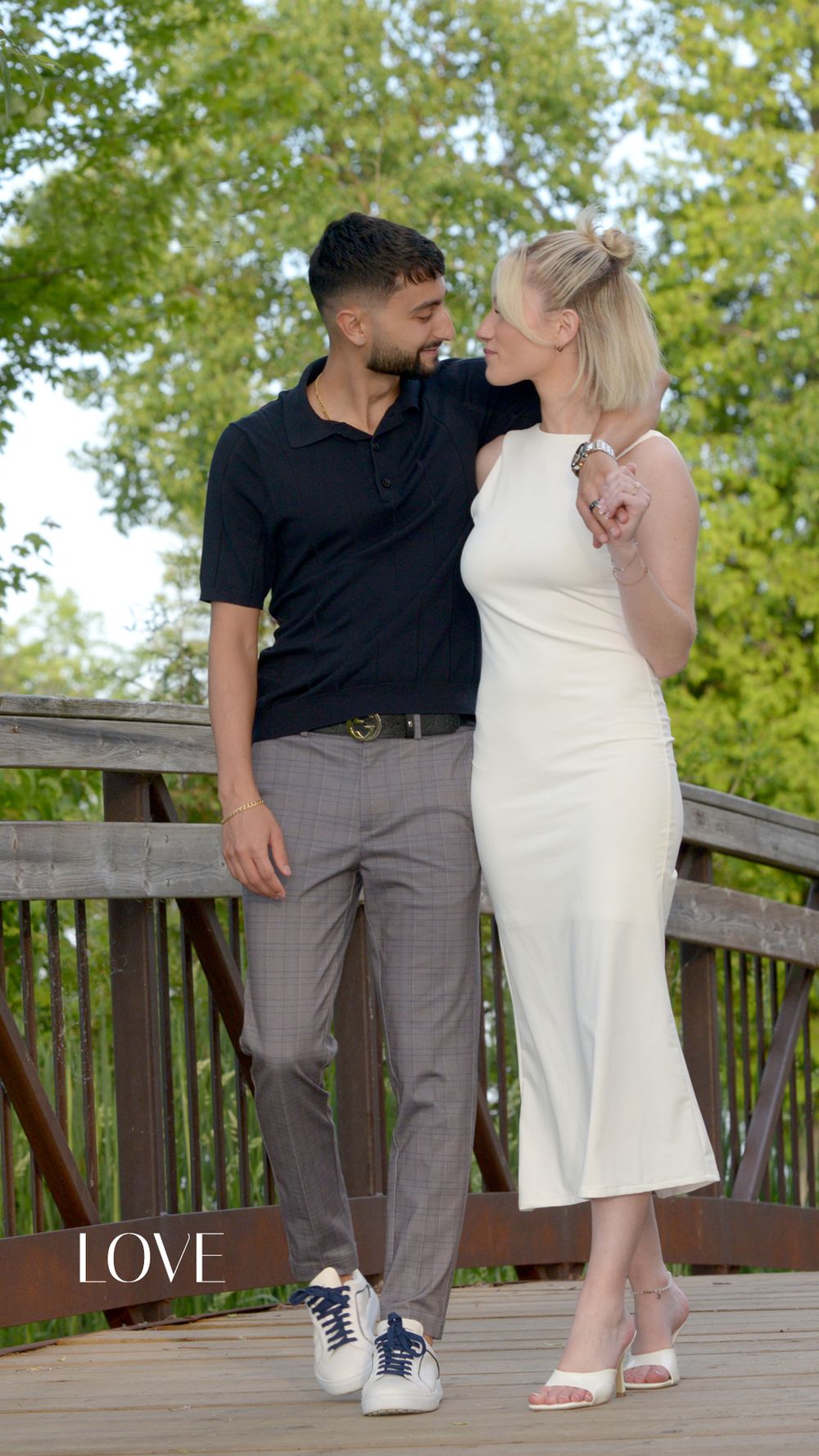 A couple shares a romantic moment on a wooden bridge surrounded by lush green trees, man in black polo and woman in white dress.
