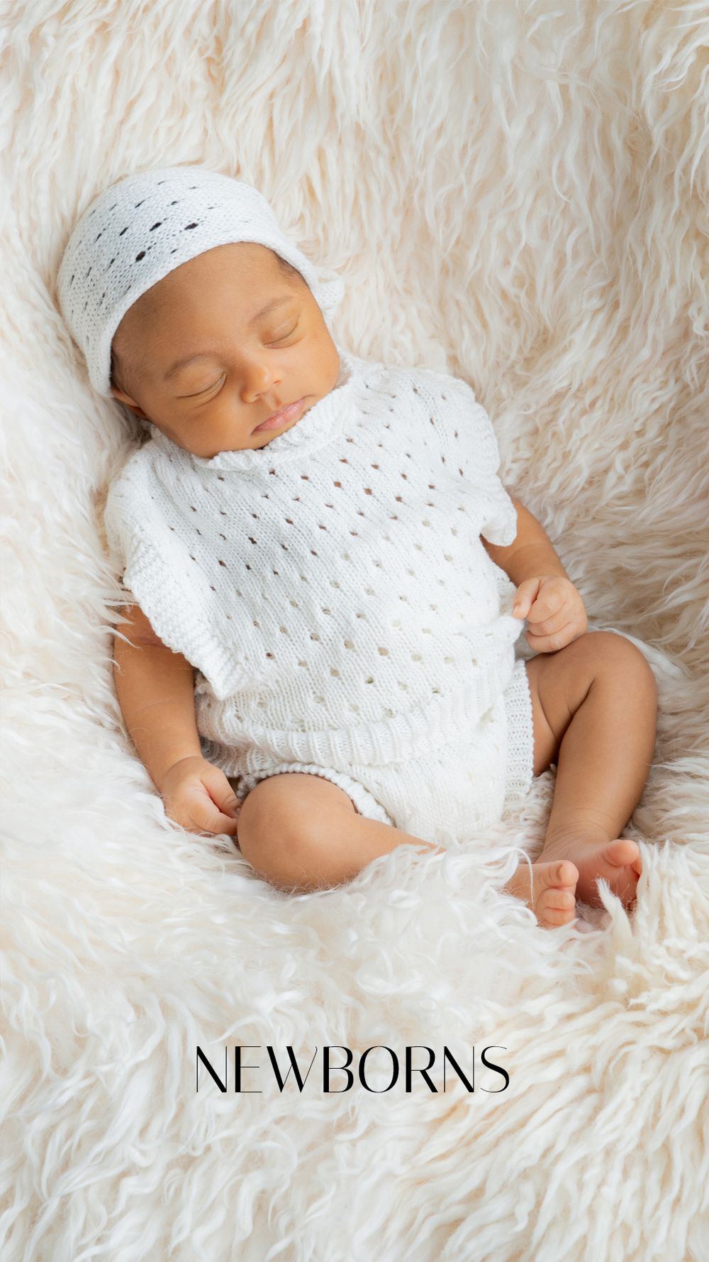 Sleeping newborn baby in white knit outfit and bonnet on fluffy cream fur backdrop.