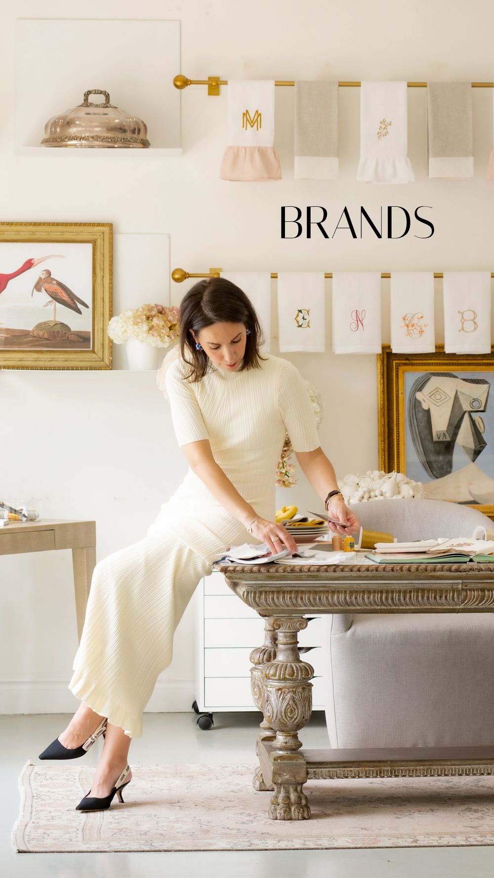 Woman in cream outfit arranging items on ornate table in elegant studio with artwork and monogrammed banners, text reads BRANDS.