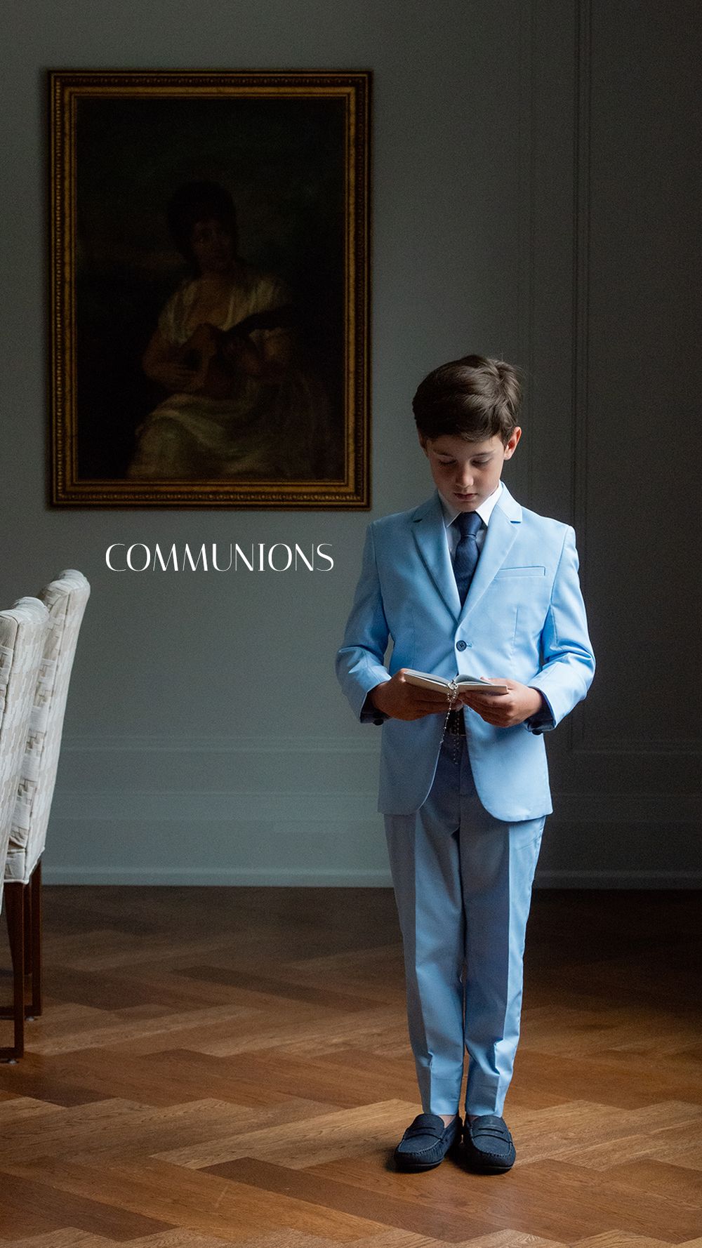 A boy in a light blue suit holds a book, standing before a classic painting, dressed for his First Communion.
