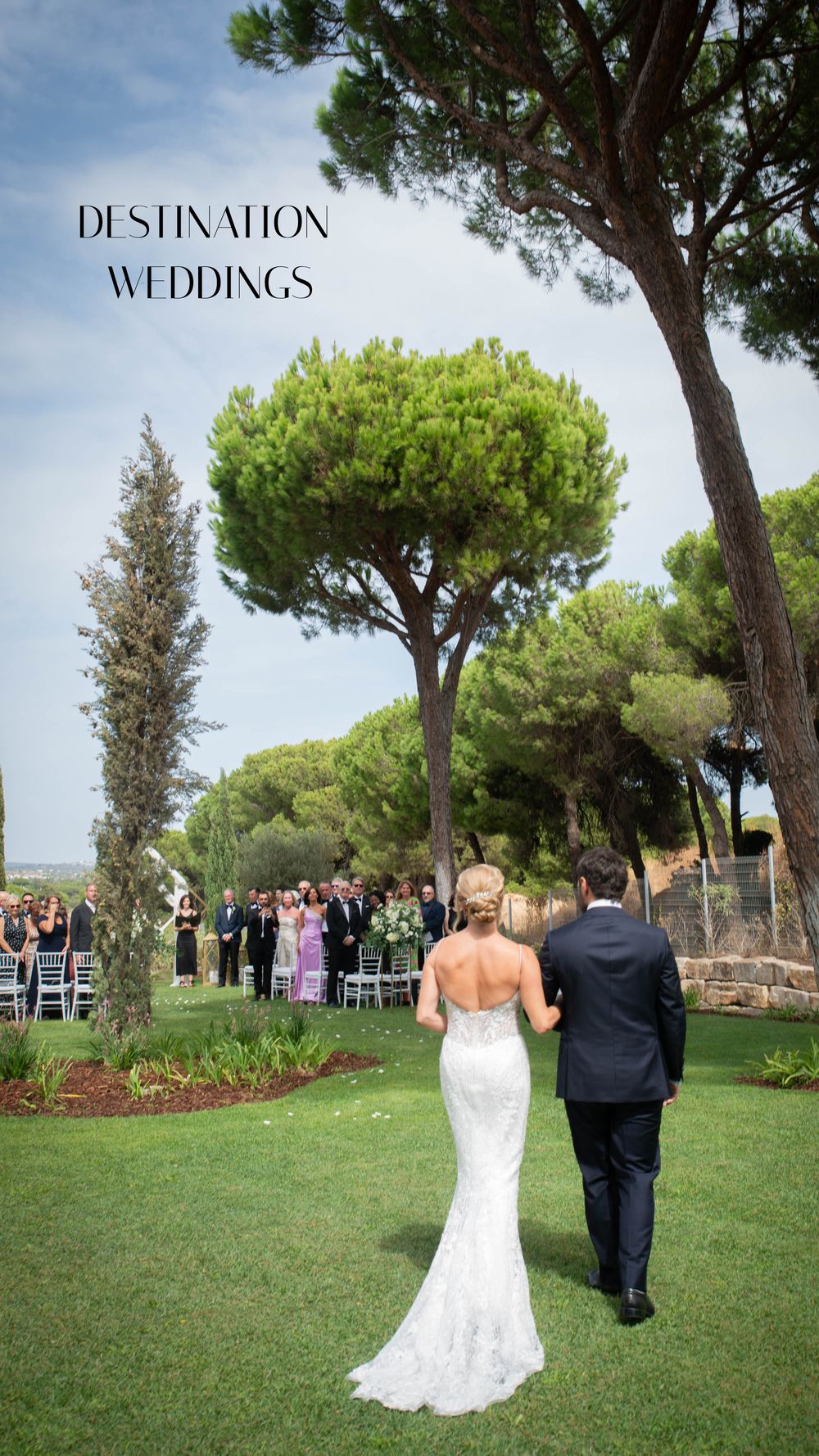 Bride and groom walk toward outdoor wedding ceremony surrounded by lush green trees at a destination wedding venue.