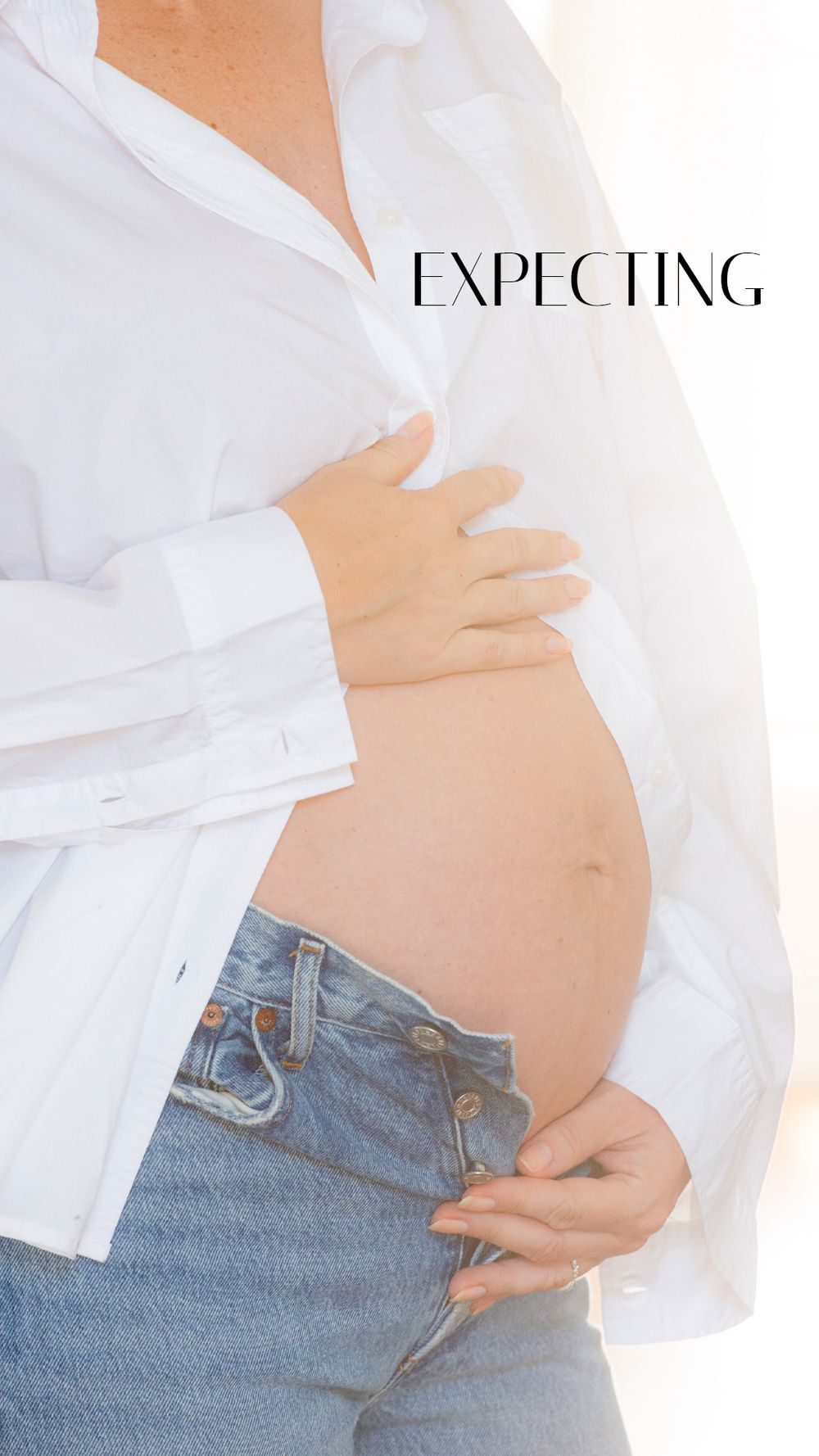 Pregnant woman in white shirt and jeans cradling baby bump with text saying Expecting.
