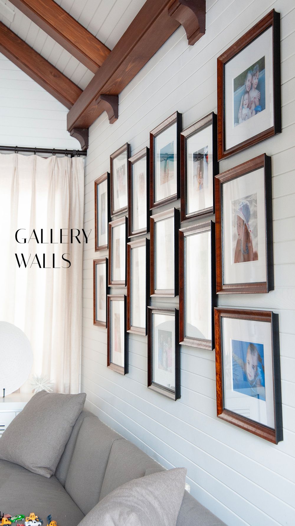 A gallery wall of wooden framed photos arranged diagonally on white shiplap wall with exposed wood beam ceiling.