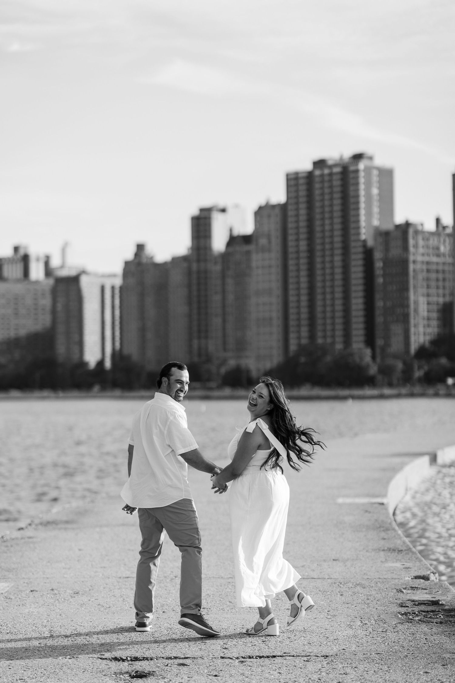 Black and white photo of a couple holding hands and walking along a lakeside path with a city skyline in the background.