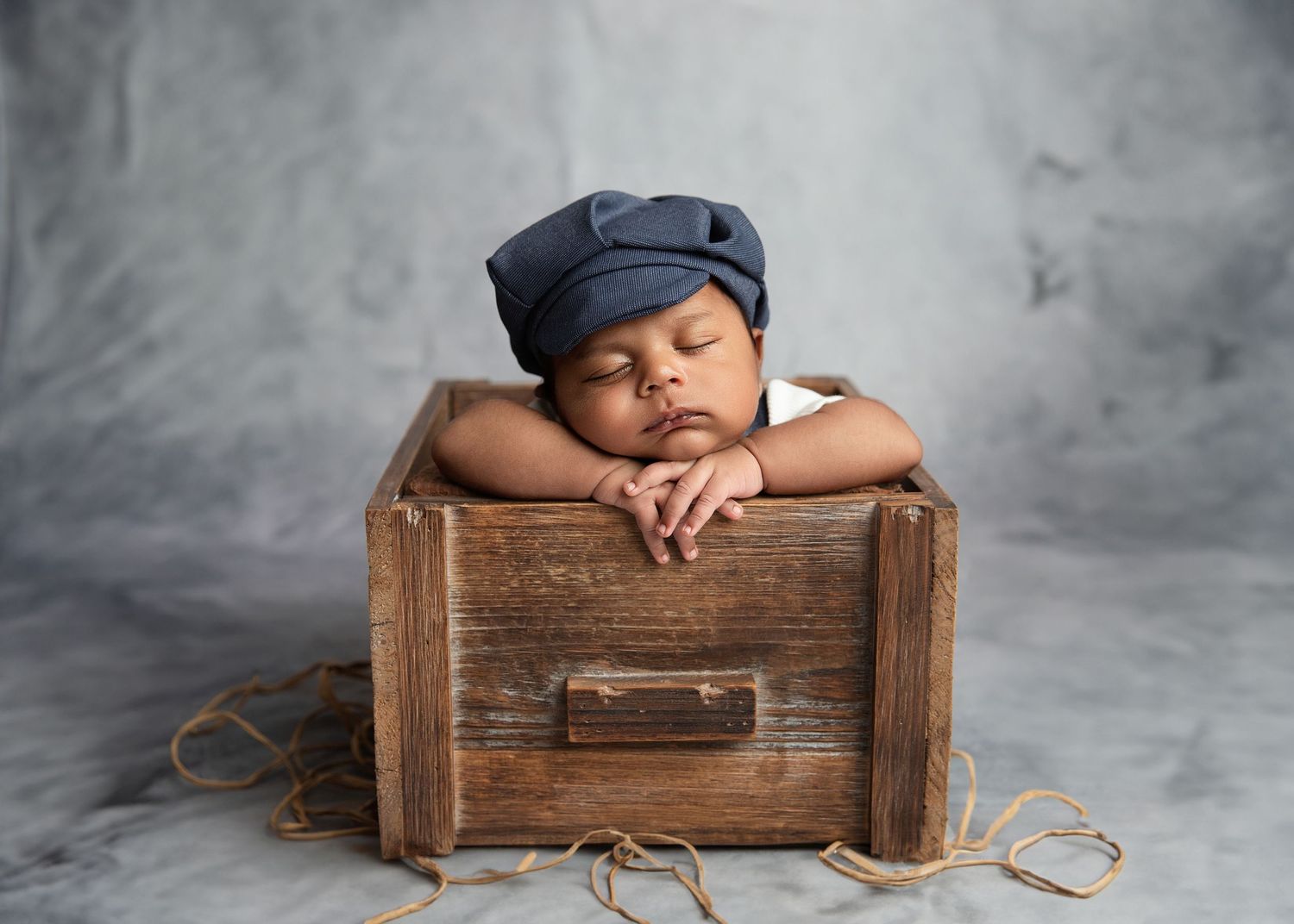 Sleeping newborn baby wearing a blue denim hat resting in a rustic wooden box on a gray background.