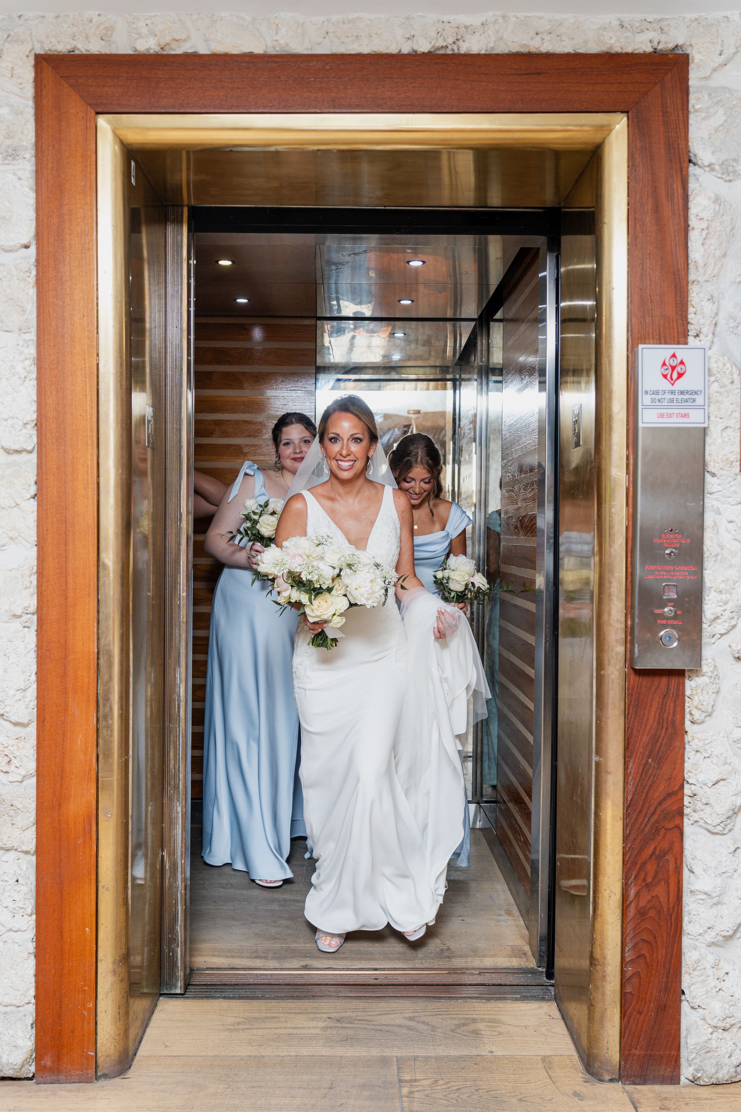 Bride and bridesmaids in blue dresses holding white bouquets stepping out of a gold elevator on wedding day.