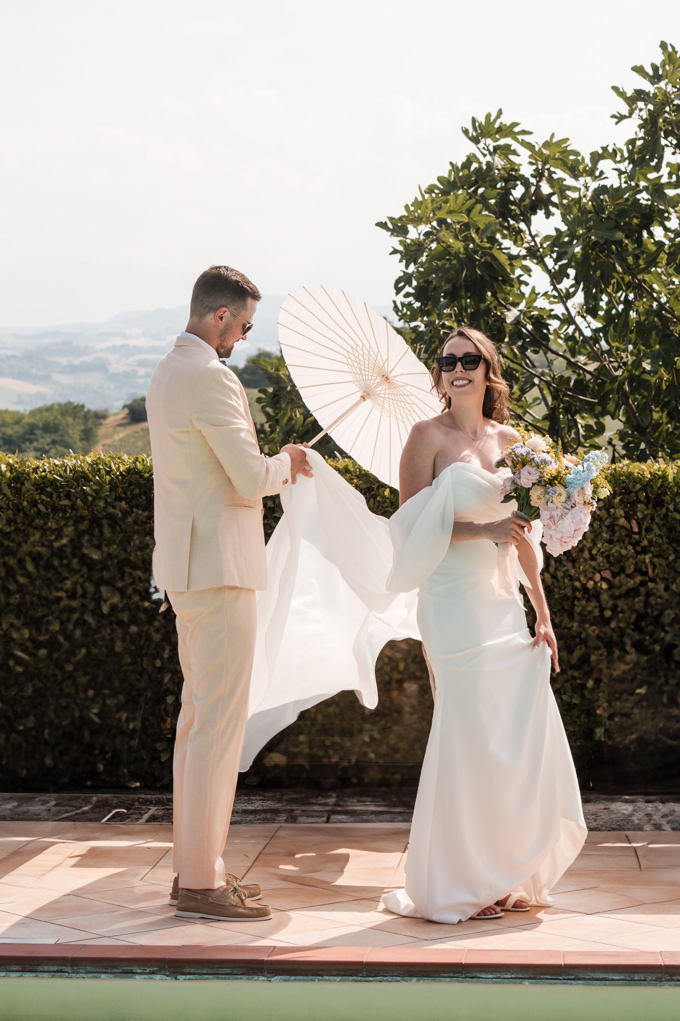 Smiling bride in white gown holds bouquet as groom holds parasol on sunny Tuscan terrace.