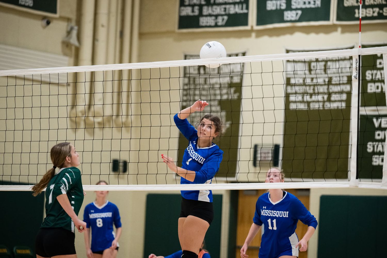 Blue-uniformed volleyball player spikes ball over net during competitive indoor high school match.