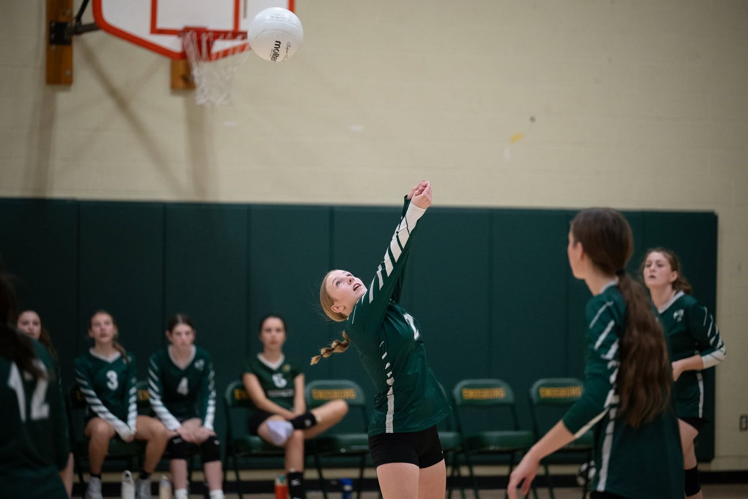 Green-uniformed volleyball player serves ball during high school match with teammates on bench.