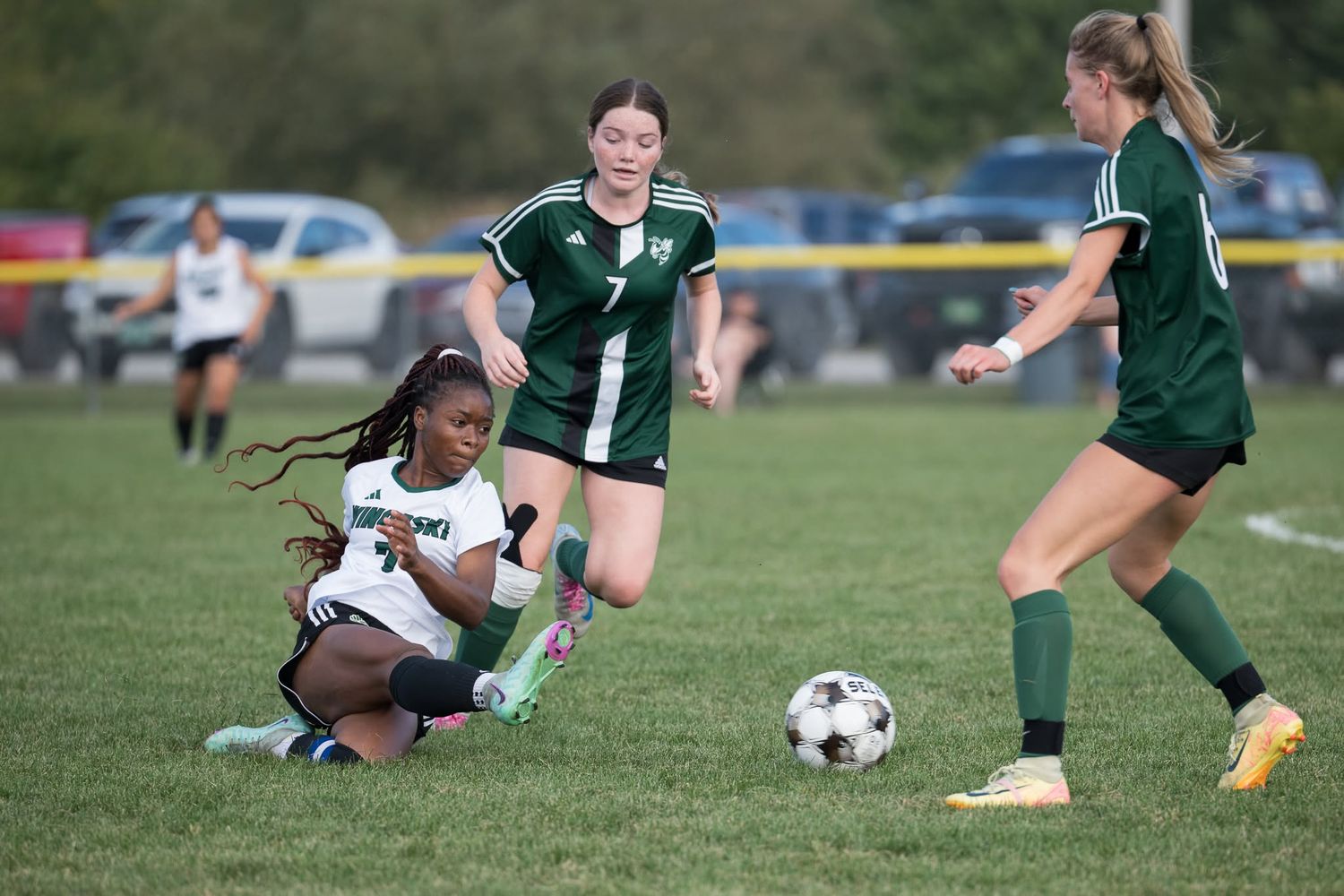 Girls soccer players compete for ball during outdoor match with green-uniformed home team.