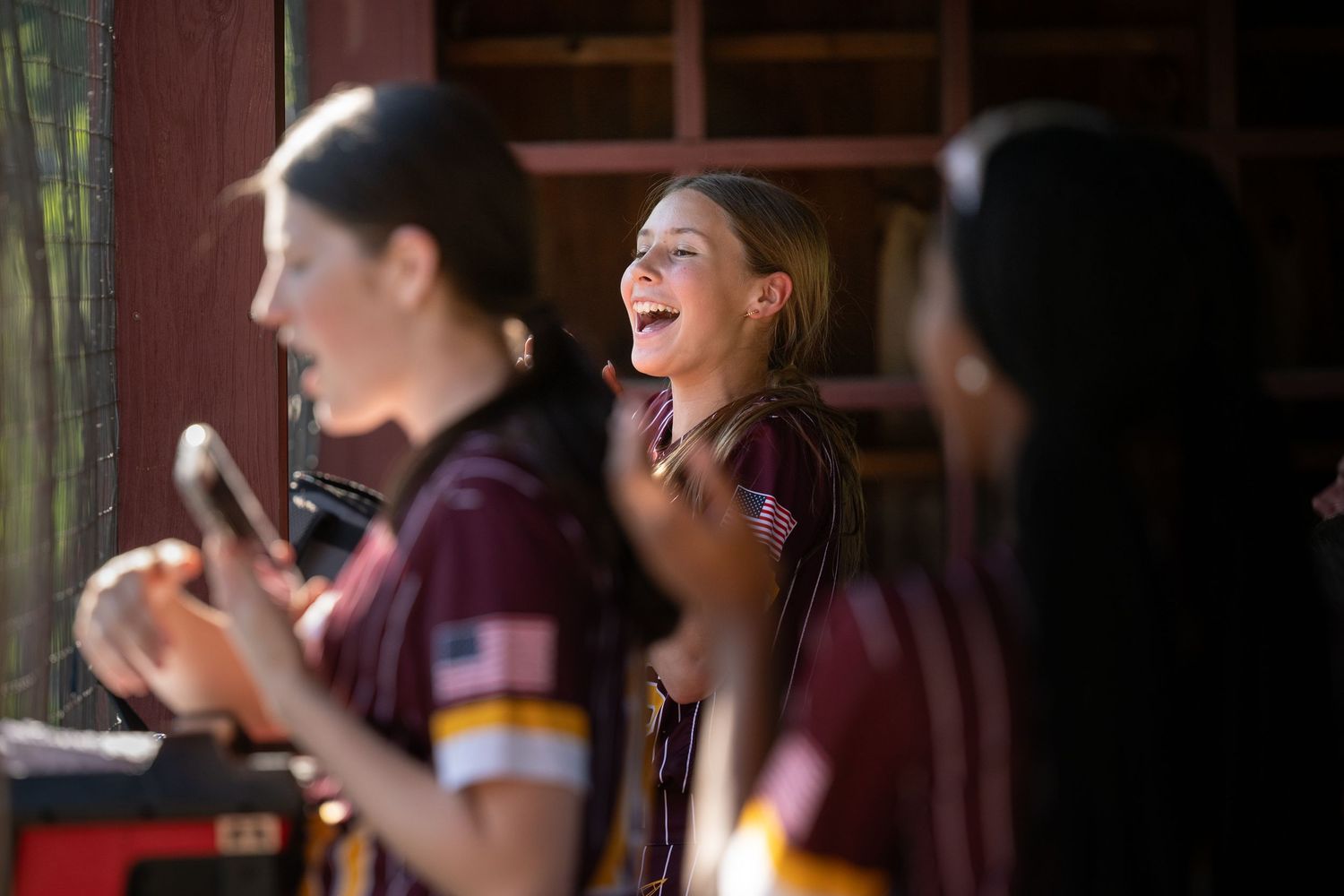 Female softball players in maroon uniforms laugh and cheer from their dugout during a game.