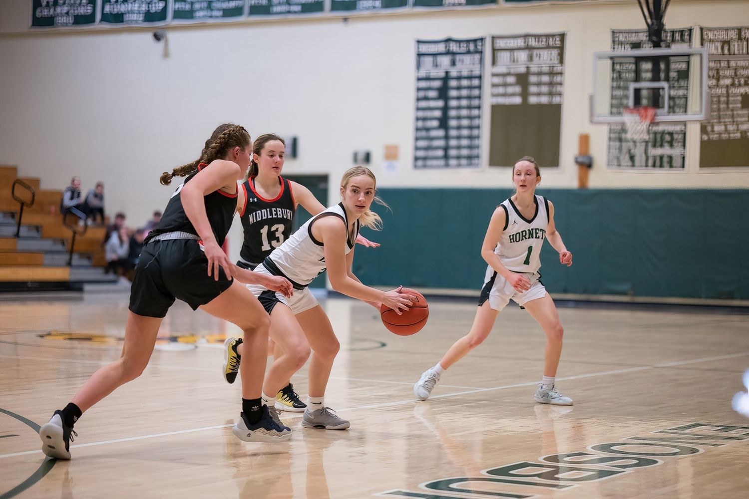 Girls basketball players compete for ball during indoor high school game with green and black uniforms.