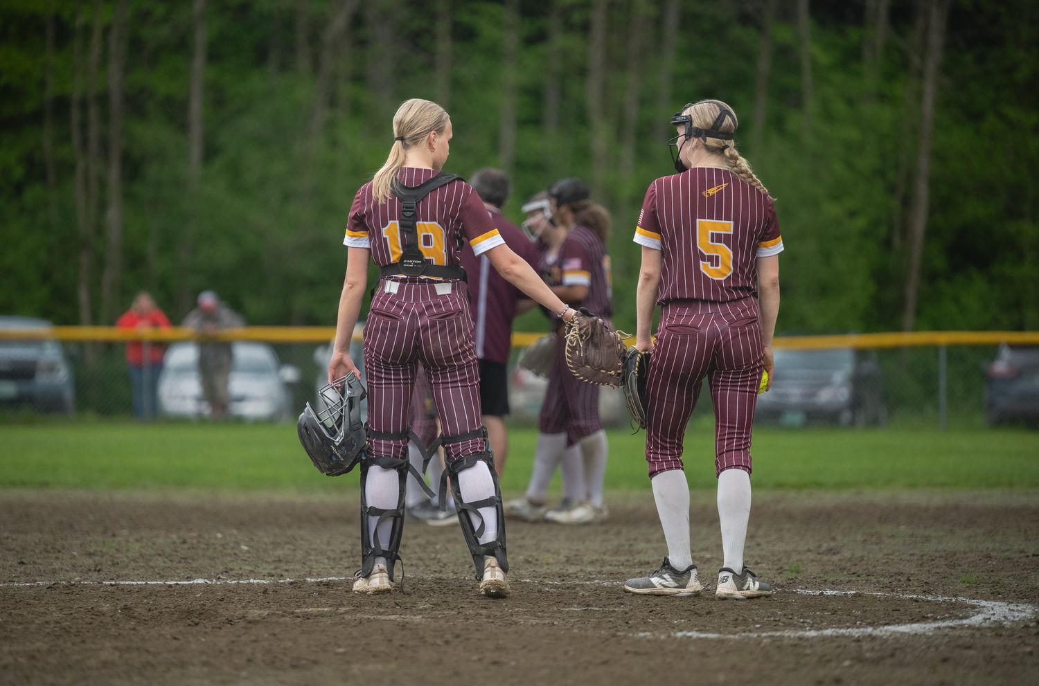 Two softball players in maroon striped uniforms stand on field, one holding catcher's gear.