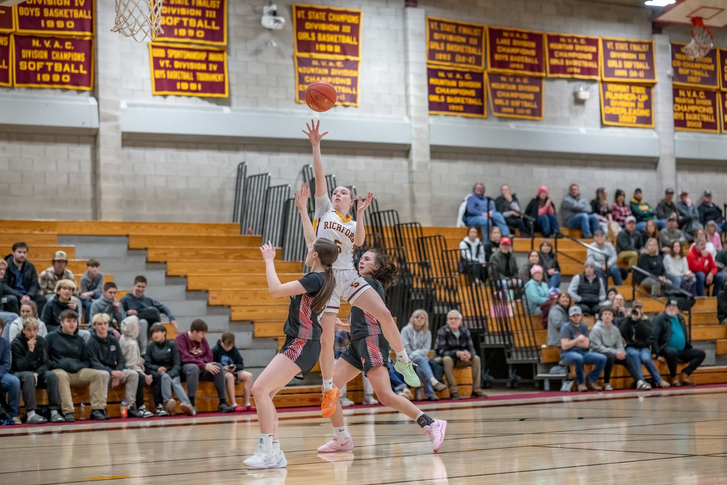Girls basketball player shoots jump shot over defender in packed high school gymnasium game.