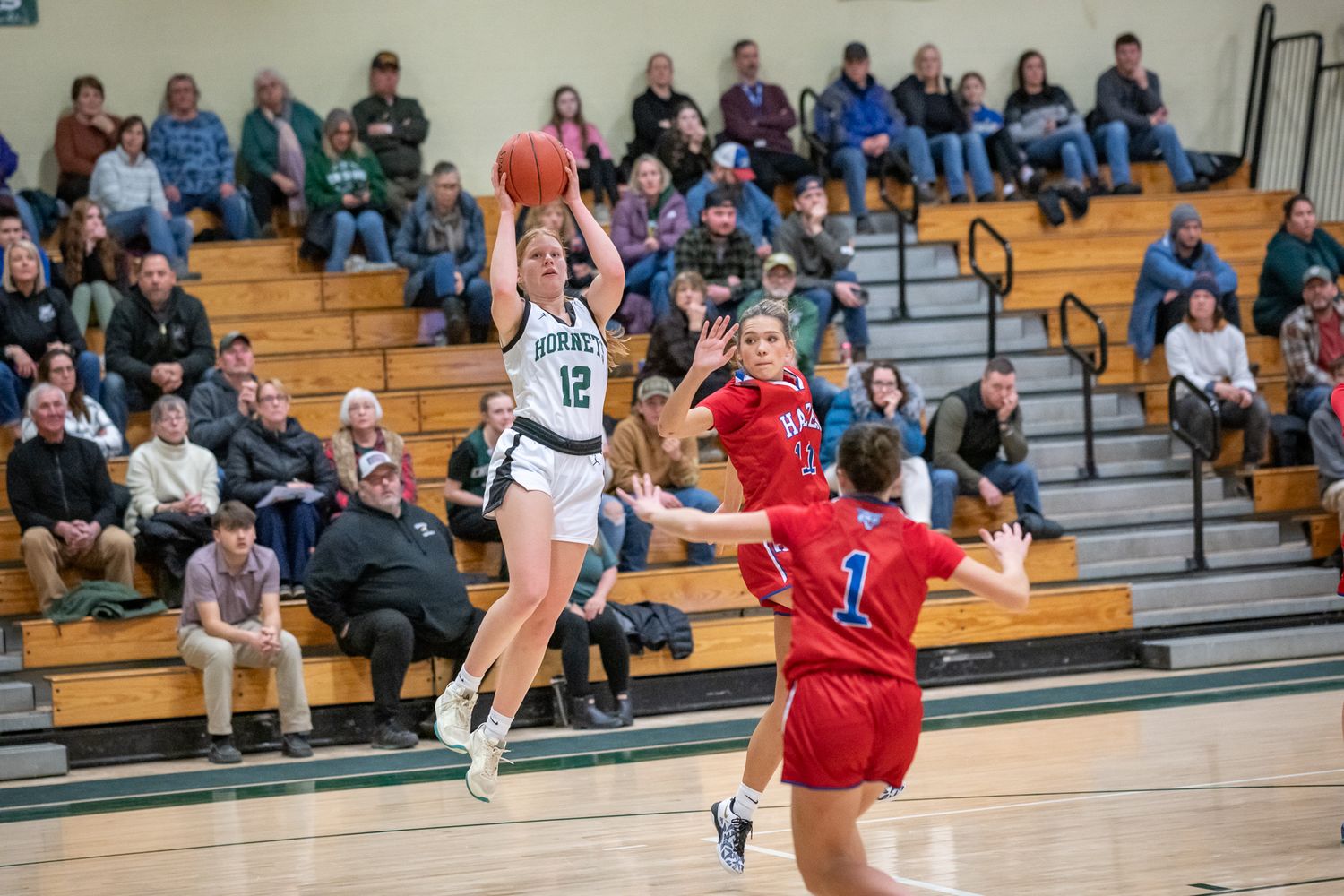 Girls basketball player #12 shoots jump shot over defenders during competitive high school game.
