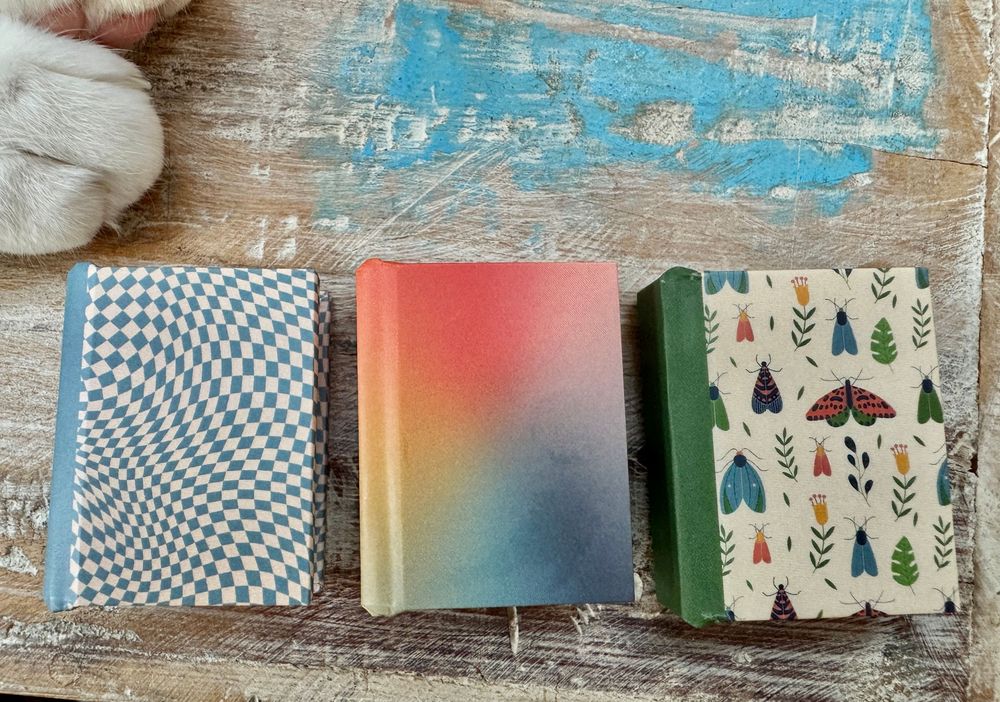 Three decorative journals with colorful covers on a rustic wooden surface, featuring checkered, gradient, and floral moth patterns.