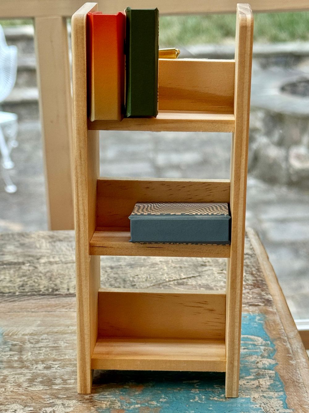 Small wooden ladder shelf with colorful books and a gray box, displayed on a rustic table near a window.