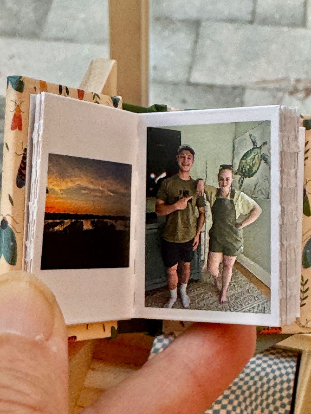 Tiny handmade miniature photo book open showing small printed photos of a sunset and two people posing indoors.