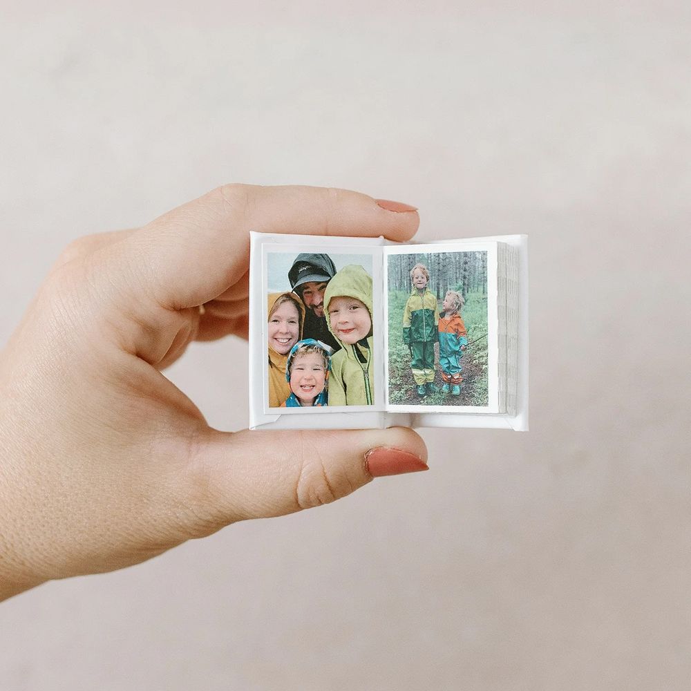 A hand holds a tiny photo album open displaying two small family photos with smiling people in outdoor clothing.