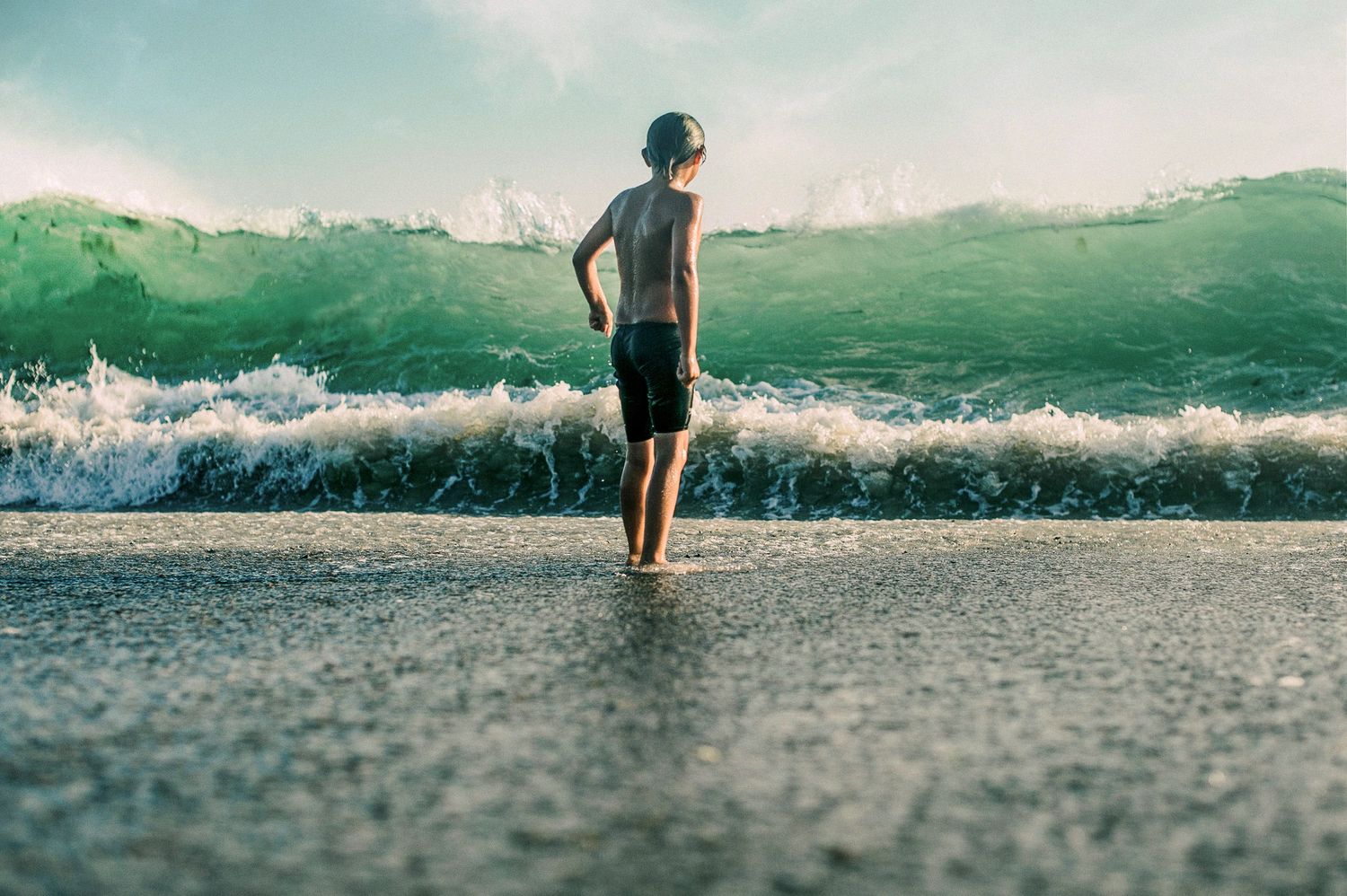 A young boy stands facing a large crashing wave on a sandy beach, wearing dark swim shorts.