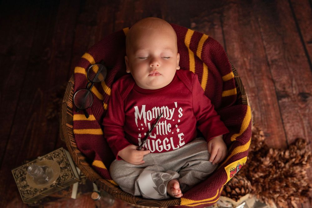 Newborn baby sleeping in basket wearing Mommy's Little Muggle onesie with Gryffindor scarf Harry Potter themed photo.