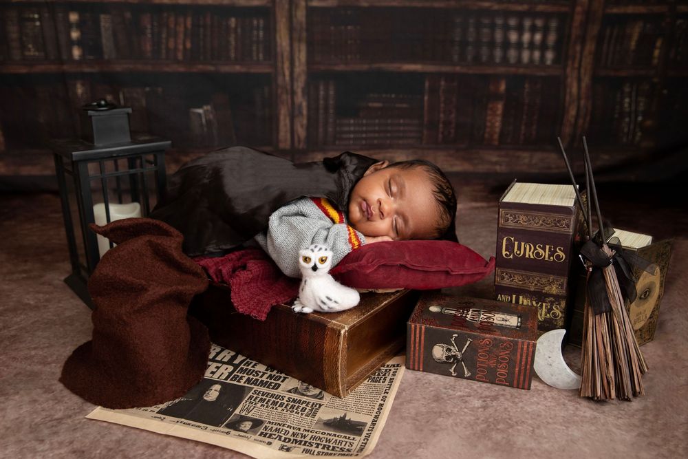Newborn baby dressed as Harry Potter sleeping among wizard props including books, a wand, and a toy owl.
