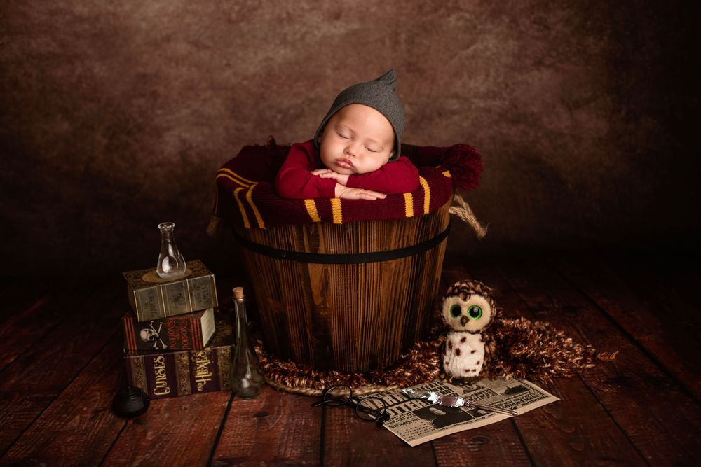 Newborn baby sleeping in wooden bucket with Harry Potter themed props, owl, and wand on rustic floor.