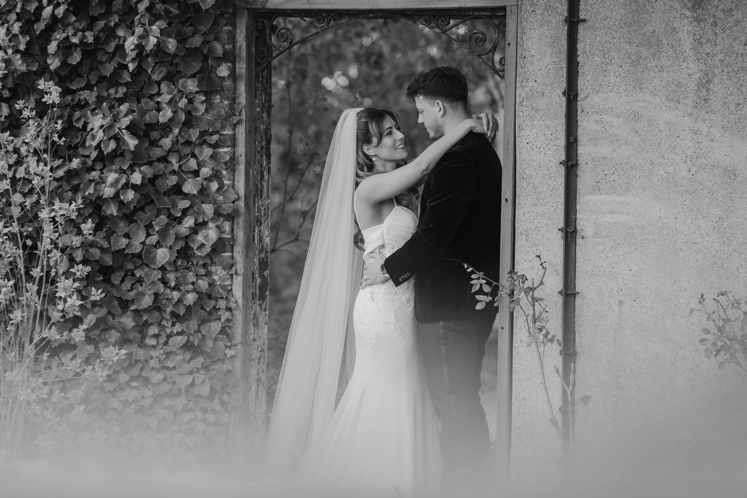 Black and white wedding photo of bride and groom embracing in a vine-covered doorway, sharing a romantic moment.
