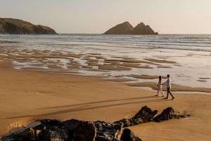 Couple walking hand in hand along a golden sandy beach at sunset with rocky islands in the distance.