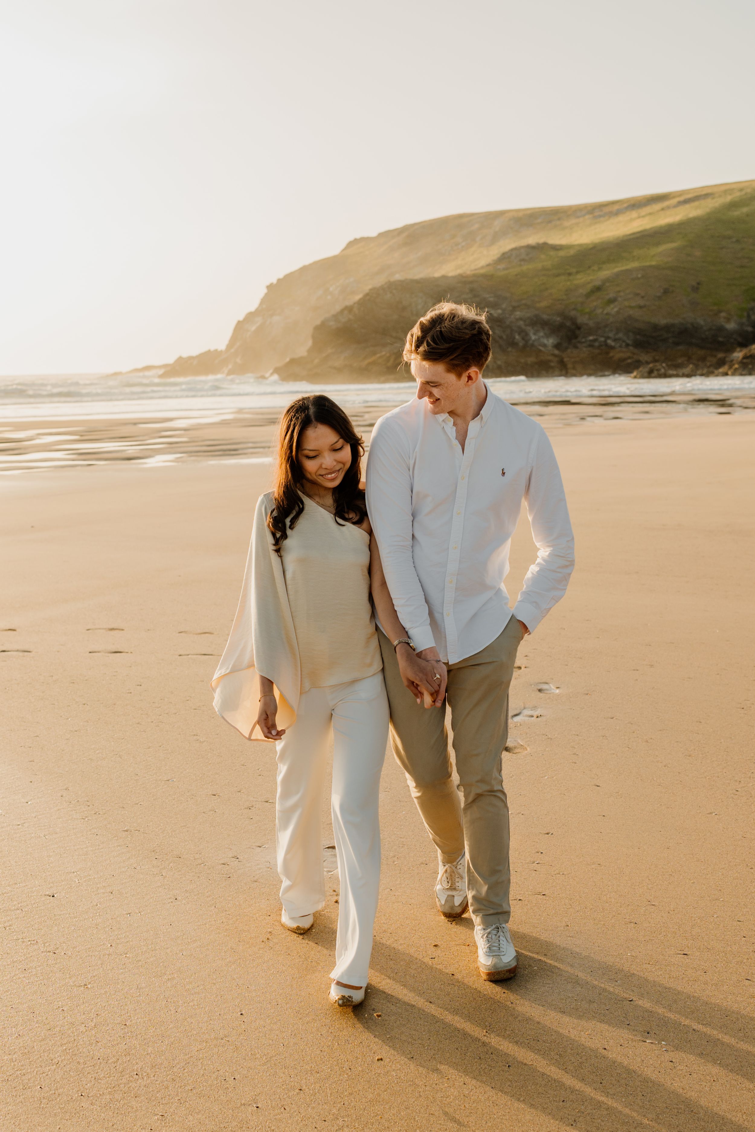 Happy couple walking hand in hand on sunlit sandy beach with green coastal cliffs in background.