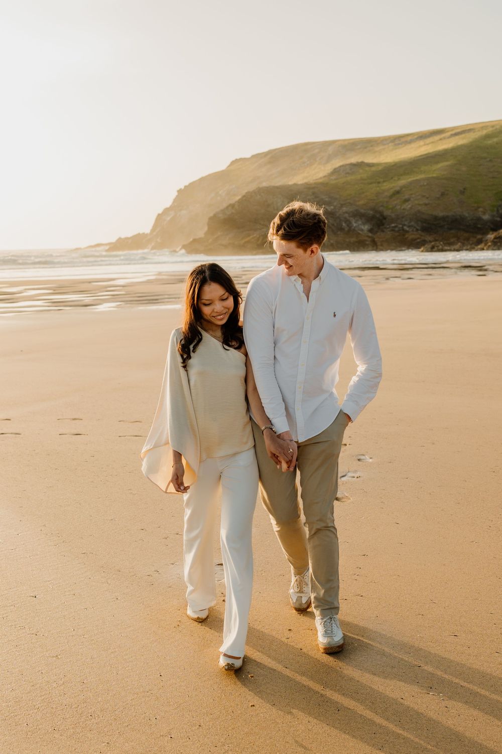 Happy couple walking hand in hand on sunlit sandy beach with green coastal cliffs in background.