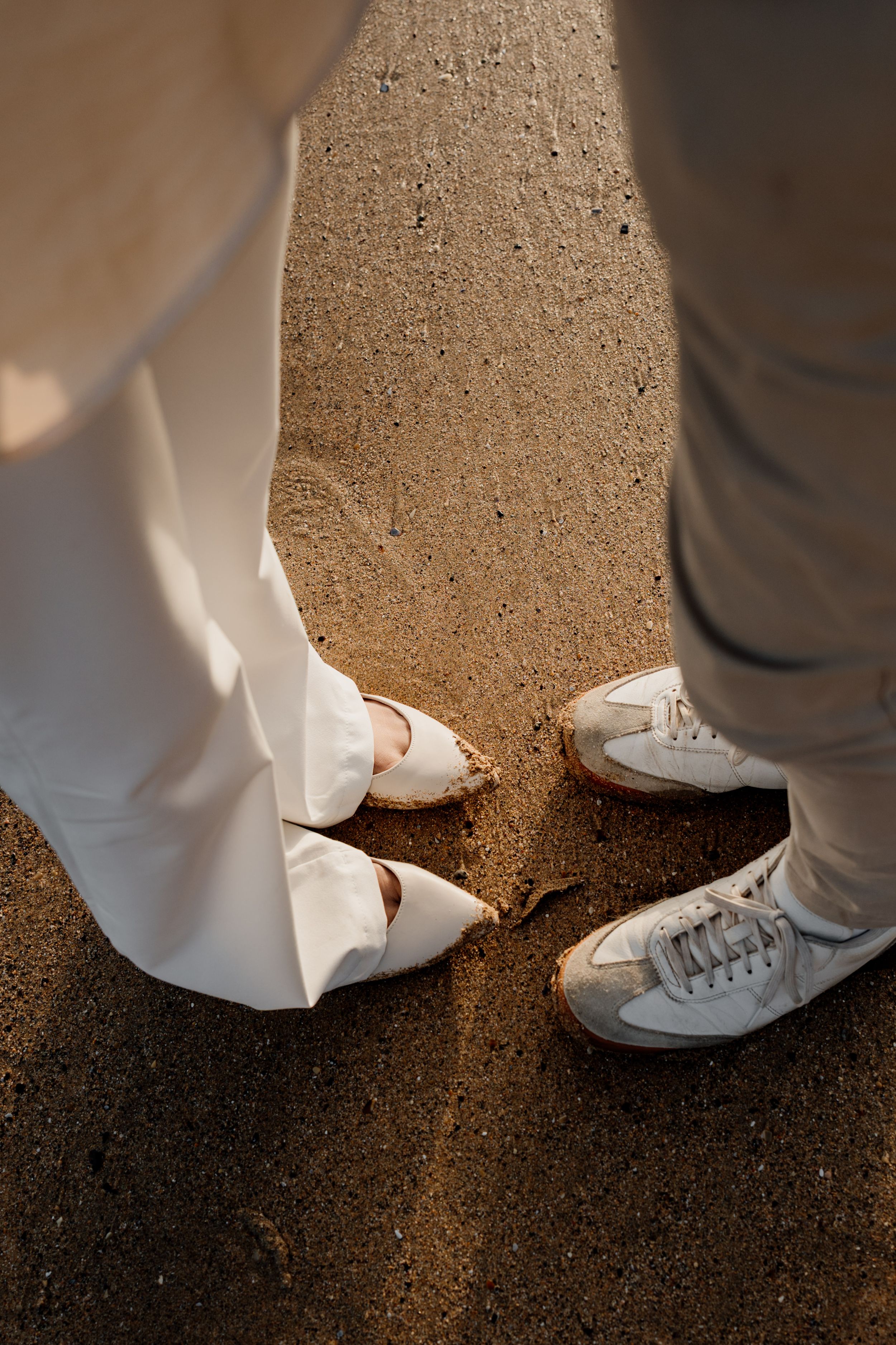 Close-up of couple's feet in white heels and sneakers standing together on golden sandy beach.