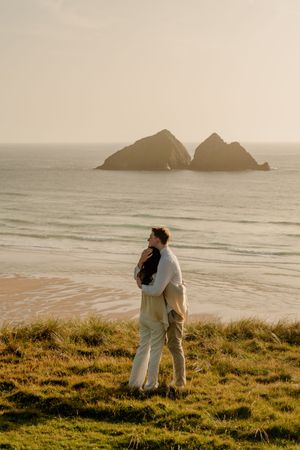 Couple embracing on grassy cliff overlooking sandy beach with twin rock formations in golden sunset light.